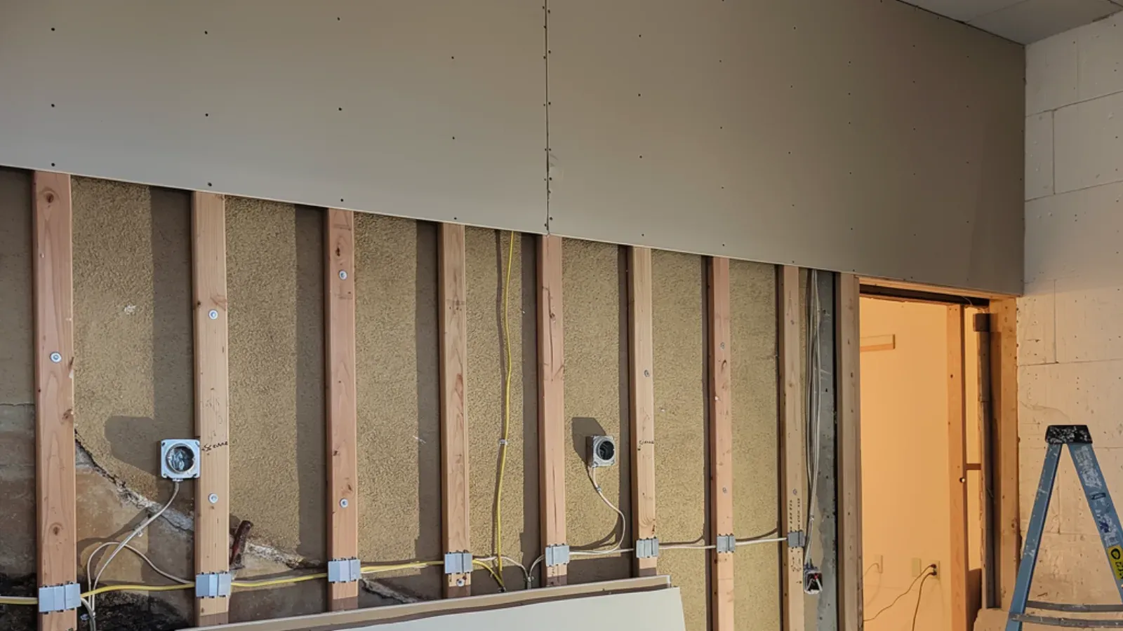Bare gypsum drywall hung across the upper half of an interior wall, the lower stud bays still open with insulation and electrical boxes visible