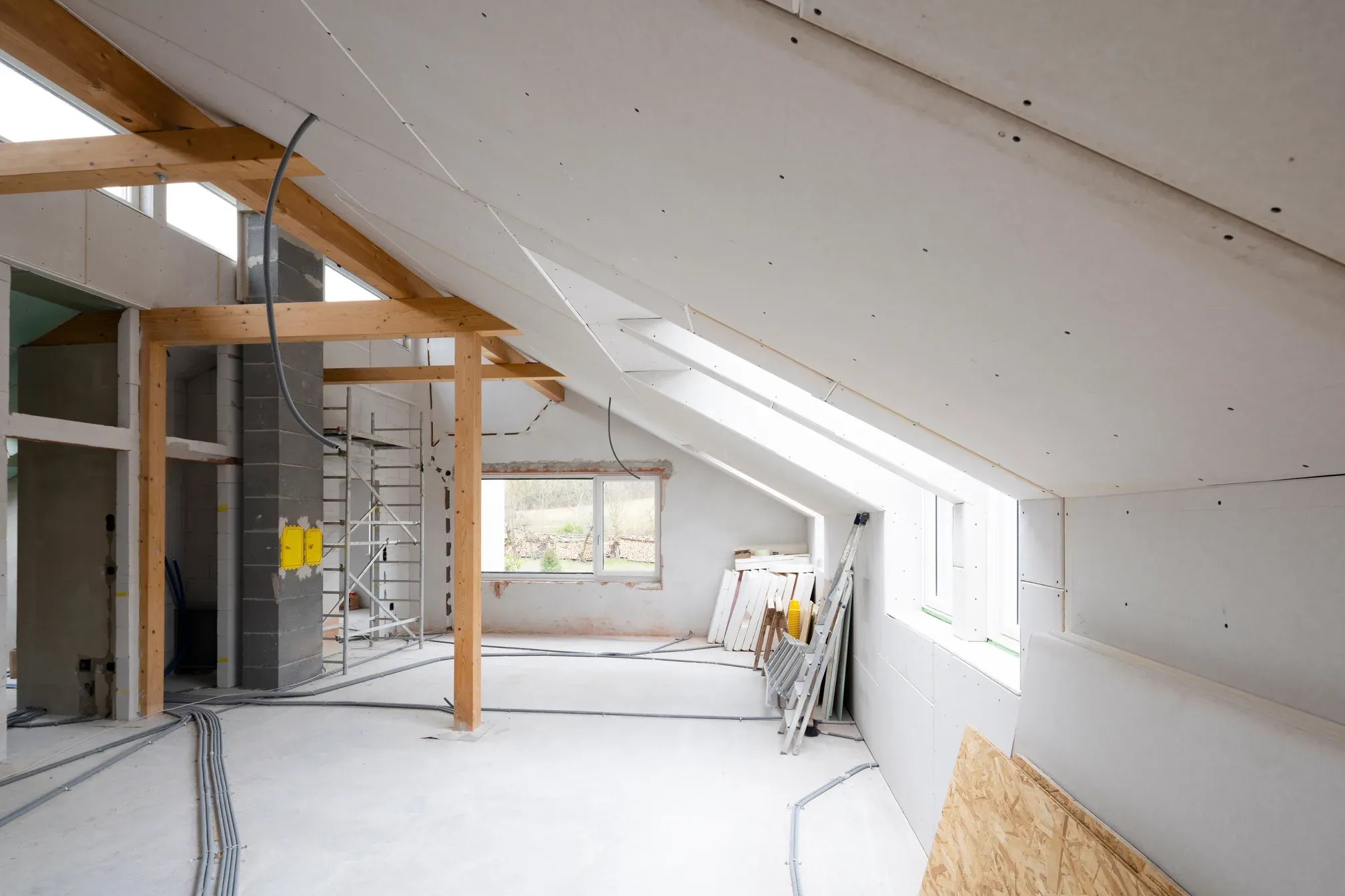 Loft under a sloped drywall ceiling with an exposed wood post-and-beam frame, hung and taped board, scaffolding, and conduit run across the bare floor
